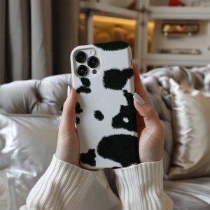 May include: A white and black cow print phone case for an iPhone. The case has a raised camera bump and a black and white cow print design.