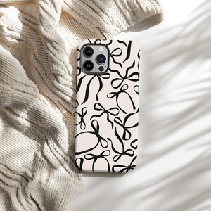 May include: A white iPhone case with a black and white pattern of bows. The case has a camera lens cutout and a black border.