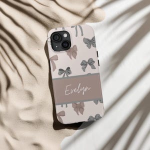 May include: A personalized phone case with a light pink background and a pattern of gray bows. The name "Evelyn" is printed in a brown script font on a gray band across the middle of the case.