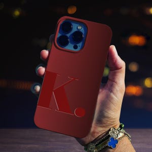 May include: A red phone case with a large white letter "K" and a red dot. The case is designed for a phone with a triple camera lens.