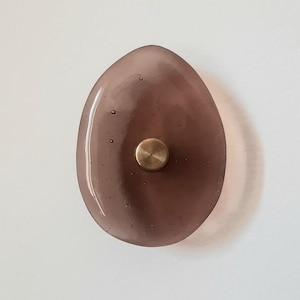 Op de afbeelding: Een druppelvormige, doorschijnende, paarse glazen kastknop met een messingkleurig metalen midden. Het glas heeft kleine luchtbellen. De knop is ontworpen voor gebruik op kasten en lades.