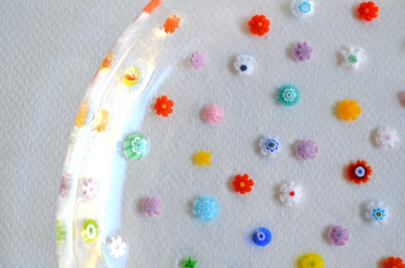 Puede incluir: Un plato de vidrio transparente adornado con numerosas flores de vidrio peque&ntilde;as y coloridas. Las flores, en tonos de rojo, azul, amarillo y blanco, son de diversos dise&ntilde;os y est&aacute;n dispersas sobre la superficie del plato.