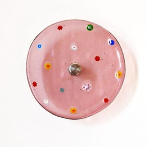 Puede incluir: Un plato de vidrio rosa con un pomo central de metal y diseños florales coloridos. El plato presenta pequeños detalles florales multicolores en rojo, amarillo, blanco, azul y verde. El plato es redondo.