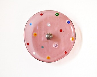Könnte beinhalten: Eine kreisförmige, rosafarbene Glasschale mit einer glatten, durchscheinenden Oberfläche, die mit kleinen, bunten, blumenförmigen Millefiori-Mustern in Rot, Gelb, Weiß, Grün und Blau verziert ist und einen kleinen, silberfarbenen Metallknopf besitzt.