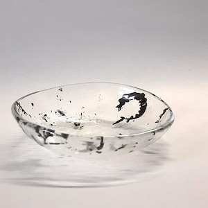 May include: A shallow, circular clear glass bowl featuring an irregular, organic rim. The interior surface is decorated with scattered black ink-like splatters and a single, prominent curved black mark resembling a stylised crescent shape.