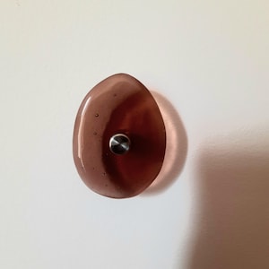 Può includere: Un gancio da parete in vetro traslucido color marrone violaceo a forma di ciottolo. Il gancio presenta una superficie liscia e arrotondata con piccole bolle d'aria interne e un elemento di fissaggio centrale in metallo color argento spazzolato.