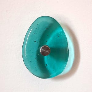 Pode incluir: Um puxador de vidro translúcido na cor verde-azulado com um formato orgânico semelhante a um seixo. A superfície do vidro apresenta pequenas bolhas de ar dispersas. Uma base circular de metal prateado escovado está centrada na peça.
