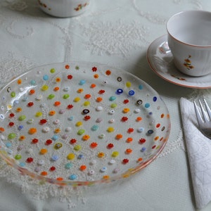 Puede incluir: Un plato de vidrio transparente decorado con diminutas cuentas de colores en forma de flor. El plato forma parte de una mesa con tazas y platillos blancos con detalles florales naranjas, un tenedor y una servilleta doblada.