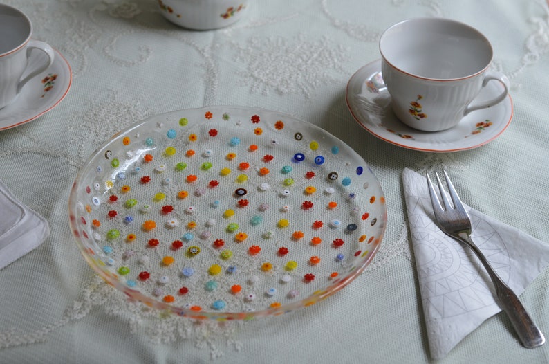 Puede incluir: Un plato de vidrio transparente decorado con diminutas cuentas de colores en forma de flor. El plato forma parte de una mesa con tazas y platillos blancos con detalles florales naranjas, un tenedor y una servilleta doblada.