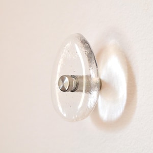 May include: Clear glass cabinet knob with a textured, oval design and a silver metal base. The knob is mounted on a white surface, casting a shadow.