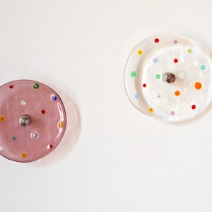 Peut inclure: Deux patères murales circulaires avec façades en verre. L'une est en verre rose translucide et l'autre en verre transparent. Les deux sont incrustées de petits motifs floraux en verre multicolore et possèdent un support central en métal argenté.