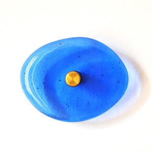 May include: A handcrafted, oval-shaped, blue glass dish with a gold-colored metal knob in the center. The glass has a glossy finish and a slightly irregular shape. The dish is approximately 4 inches in length.