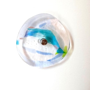 Può includere: Un piatto decorativo rotondo in vetro trasparente con accenti blu, turchese, rosa e verdi. Il piatto ha un centro in metallo ed è montato su una parete bianca. Il vetro ha un aspetto strutturato.