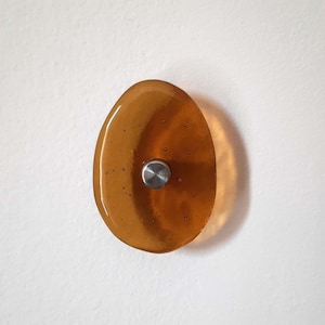 May include: An amber-coloured glass knob with a stainless steel centre. The knob is oval-shaped and has a smooth, glossy surface. The glass has a gradient effect, transitioning from a lighter to a darker amber hue.