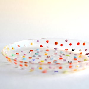 Puede incluir: Un plato de vidrio transparente y poco profundo decorado con peque&ntilde;os dise&ntilde;os florales y de puntos de colores. El plato tiene un borde ligeramente elevado y est&aacute; lleno de acentos de varios colores, incluyendo rojo, naranja, amarillo, verde, azul y morado. El plato est&aacute; sobre una superficie blanca.