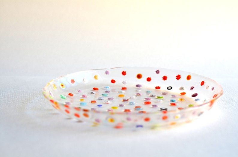 Puede incluir: Un plato de vidrio transparente y poco profundo decorado con peque&ntilde;os dise&ntilde;os florales y de puntos de colores. El plato tiene un borde ligeramente elevado y est&aacute; lleno de acentos de varios colores, incluyendo rojo, naranja, amarillo, verde, azul y morado. El plato est&aacute; sobre una superficie blanca.