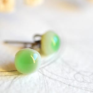 May include: A pair of stud earrings with round, light green, iridescent glass beads. The earrings are set on silver posts and are displayed on a white surface with faint, abstract markings. The earrings are in soft focus.