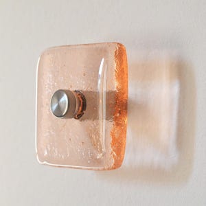 May include: A square, pale pink glass wall sconce with a brushed metal knob. The edges of the glass have a textured, amber-coloured finish. The sconce is mounted on a white wall.