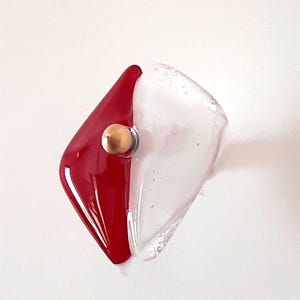 Könnte beinhalten: Ein Ring mit geteiltem Design, bei dem eine Seite aus rotem Glas und die andere aus klarem Glas besteht. Eine kleine, polierte goldfarbene Metallkugel ist in der Mitte eingelassen, wo die beiden Glasabschnitte aufeinandertreffen.