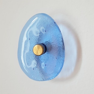 Pode incluir: Um puxador de vidro azul claro em forma de gota com uma base de metal cor de latão. O vidro tem pequenas bolhas de ar e é montado numa superfície branca. O puxador é um elemento decorativo.
