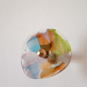 May include: A colorful glass cabinet knob with a copper-colored metal base. The knob features a clear, rounded shape with embedded pieces of blue, orange, yellow, and green glass. The glass has a textured appearance.