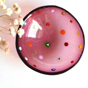 Könnte beinhalten: Eine dekorative, runde, rosa Glasschale mit dunklem Rand. Die Schale ist mit bunten, blütenförmigen Akzenten verziert. Zarte weiße Blüten befinden sich in der oberen linken Ecke und verleihen der Komposition Eleganz.