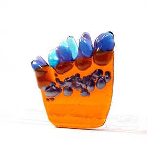 May include: Orange glass hand sculpture with blue and purple accents. The hand is textured with small bubbles and has a raised design on the fingers.