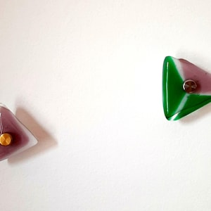 Puede incluir: Dos tiradores triangulares de cristal para armarios. Uno está dividido en secciones verdes y rosas con un tornillo plateado. El otro es rosa con un tornillo dorado. Ambos están montados sobre una superficie blanca.