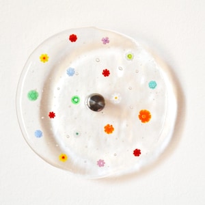 May include: Clear glass plate with colorful floral designs in red, orange, yellow, green, blue, and purple. The plate has a silver metal knob in the center. The plate is round and has a slightly irregular shape.