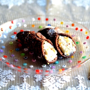 Puede incluir: Dos cannoli de chocolate rellenos de cremosa ricotta blanca, presentados en un plato de cristal transparente decorado con lunares de colores. Un tenedor plateado descansa junto al plato, sugiriendo un postre.