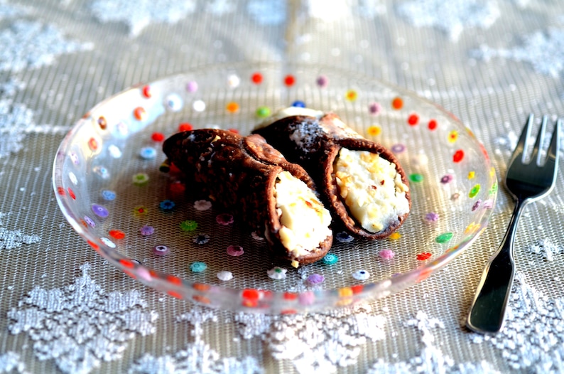 Puede incluir: Dos cannoli de chocolate rellenos de cremosa ricotta blanca, presentados en un plato de cristal transparente decorado con lunares de colores. Un tenedor plateado descansa junto al plato, sugiriendo un postre.