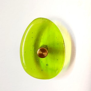 Puede incluir: Un pomo de armario de vidrio verde lima con un centro color latón. El pomo tiene forma ovalada con pequeñas burbujas de aire visibles en el vidrio. El pomo está montado sobre una superficie blanca.