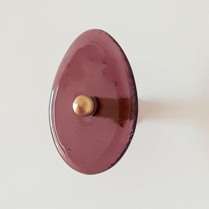 以下が含まれることがあります： 光沢のある楕円形の紫色のガラス製家具用ノブ。表面は滑らかで反射し、小さな気泡が含まれています。紫色のガラスの前面中央には、小さな金色の金属製円形スタッドが取り付けられています。
