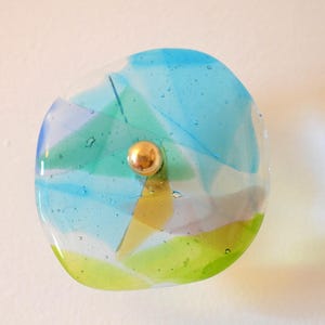 May include: A round, colorful glass wall sconce with a gold-colored center. The glass is a mix of blue, green, and yellow, with a textured, layered appearance. The sconce is mounted on a white wall.