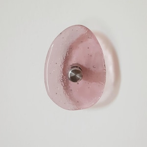 May include: A pink, teardrop-shaped glass wall hook with a stainless steel knob. The glass has a textured surface with small air bubbles. The hook is mounted on a white wall.