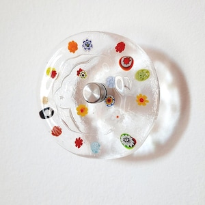 May include: A clear glass wall sconce with colorful floral designs. The sconce has a silver metal center and is decorated with red, yellow, orange, and blue flower patterns. The glass is round and has a slight curve.