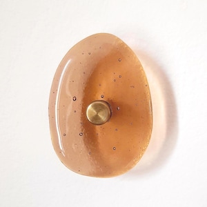 Op de afbeelding: Een ovale wandhaak met een doorschijnend, amberkleurig harslichaam met overal zichtbare luchtbellen. Een kleine, ronde metalen knop in geborstelde messingkleur is gecentreerd op het vooroppervlak van het harsstuk voor functioneel gebruik.