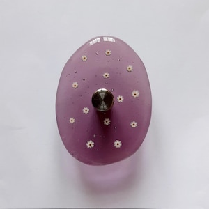 May include: An oval-shaped purple resin drawer knob featuring embedded small white daisy flowers with yellow centers. A polished silver-toned metal base is centered on the front of the translucent purple resin surface, which also contains tiny air bubbles.