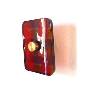 May include: A rectangular wall hook featuring a glossy, fused glass plate with a red, orange, and purple plaid pattern. The plate is mounted with a central, circular, brushed gold-toned metal knob that protrudes from the front surface.