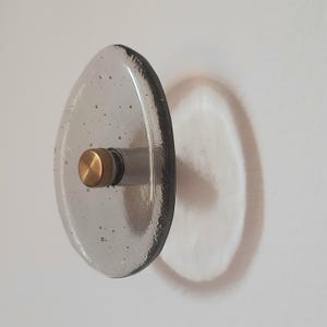 Puede incluir: Un pomo de vidrio transparente de forma ovalada con una base de metal color latón. El pomo está montado en una pared blanca, proyectando una sombra. El vidrio tiene pequeñas burbujas, agregando textura. Adecuado para armarios o cajones.