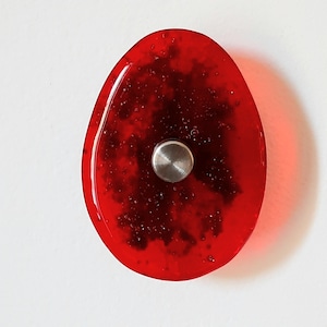 May include: A translucent, oval-shaped red glass cabinet knob featuring a smooth, glossy surface with embedded dark speckles and a central circular brushed metal attachment point. The design is simple and functional, showcasing a vibrant red hue.