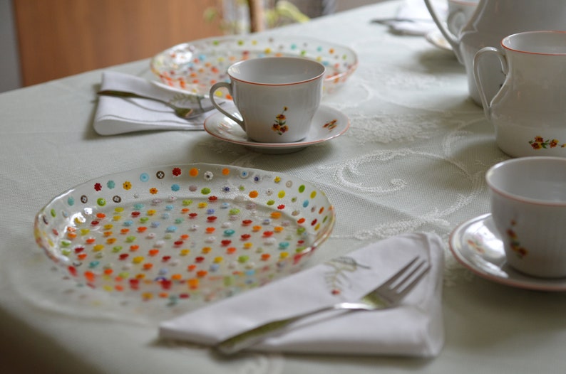 Puede incluir: Una mesa con platos de vidrio transparente adornados con dise&ntilde;os florales coloridos. Tazas y platillos blancos con detalles florales y una tetera blanca tambi&eacute;n est&aacute;n presentes. Servilletas y cubiertos completan la elegante disposici&oacute;n.