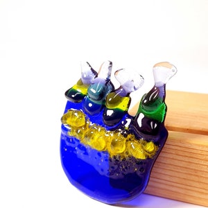 May include: A blue glass sculpture with three figures, each with a different color scheme of green, yellow, and purple. The figures are arranged in a cluster and are set against a wooden background.