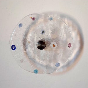 Puede incluir: Un pomo de armario redondo de cristal transparente con base de metal plateado. El cristal está decorado con pequeños diseños florales de colores. El pomo está montado sobre una superficie blanca, proyectando una sombra.