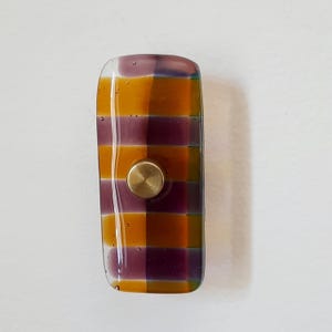 May include: A rectangular glass cabinet knob featuring a grid pattern of translucent purple and amber squares. The knob has a smooth, glossy finish with small air bubbles visible within the glass and a centered, circular brushed brass metal base.