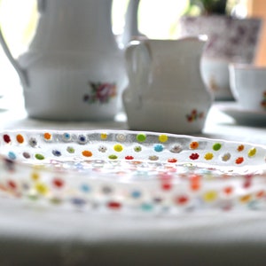 Puede incluir: Una fuente ovalada de vidrio transparente con dise&ntilde;os florales punteados de colores. Tazas de t&eacute;, platillos y una jarra de cer&aacute;mica blanca en el fondo, sugieren un ambiente de merienda.