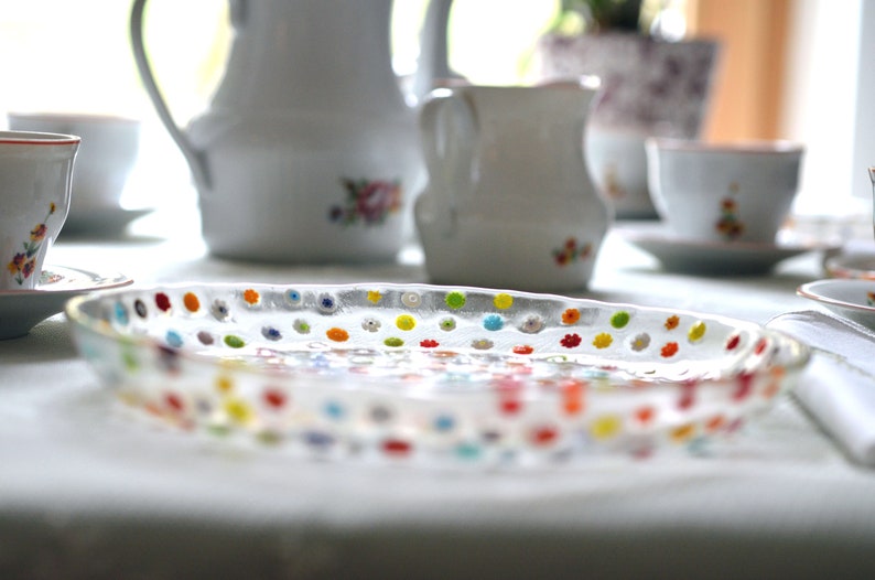 Puede incluir: Una fuente ovalada de vidrio transparente con dise&ntilde;os florales punteados de colores. Tazas de t&eacute;, platillos y una jarra de cer&aacute;mica blanca en el fondo, sugieren un ambiente de merienda.