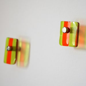 Può includere: Due ganci in vetro verde e arancione con accenti in metallo argentato. I ganci sono montati su una parete bianca.