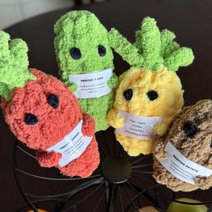 May include: Four plush toys shaped like a carrot, pickle, pineapple, and potato. Each toy has a friendly face and a small tag with a positive message. The carrot tag reads "Positive Carrot", the pickle tag reads "Positive P-ickle", the pineapple tag reads "Fine - Apple. It's fine. You're fine. Everything is fine", and the potato tag reads "Positive Potato. I may be a tiny potato, but I believe in you."