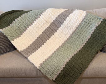 May include: A handmade, chunky knit blanket in a diagonal striped pattern. The blanket features stripes of white, gray, and various shades of green. The soft, textured blanket is draped over a gray sofa, offering a cozy and inviting aesthetic.